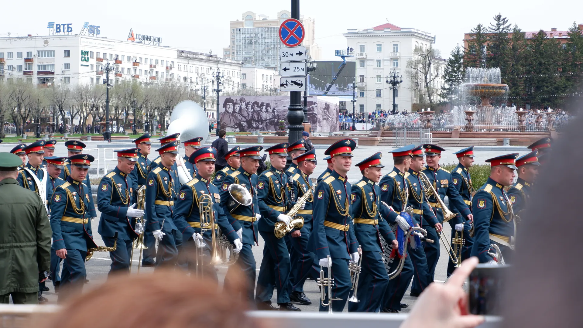 The members of the military band are holding various instruments, including a tuba and trumpet. They are dressed in uniform dark outfits and marching in sync. In the background, there is a fountain and buildings.