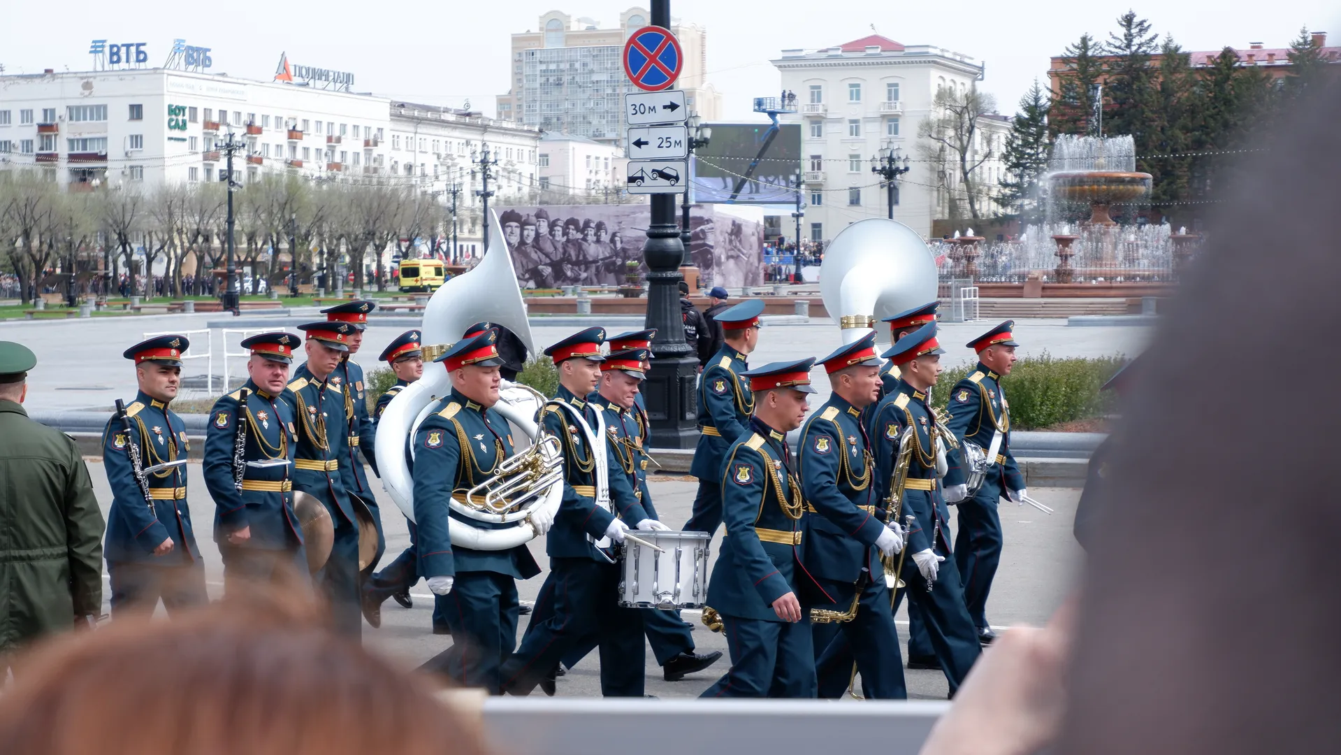 The military band continues marching, with one member holding a drum. They are wearing dark uniforms, and in the background, there are a fountain and buildings.
