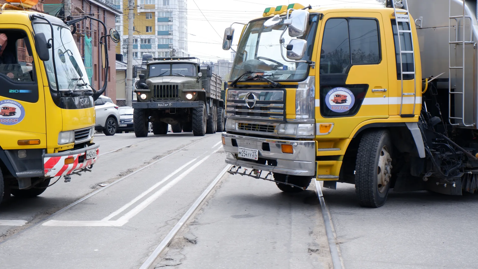 Three large trucks are parked on the street, with a military truck in the middle. The truck on the left has a “ВОСТОК” logo on the front, and the truck on the right has a round pattern on the door. The street has tracks and traffic signs.