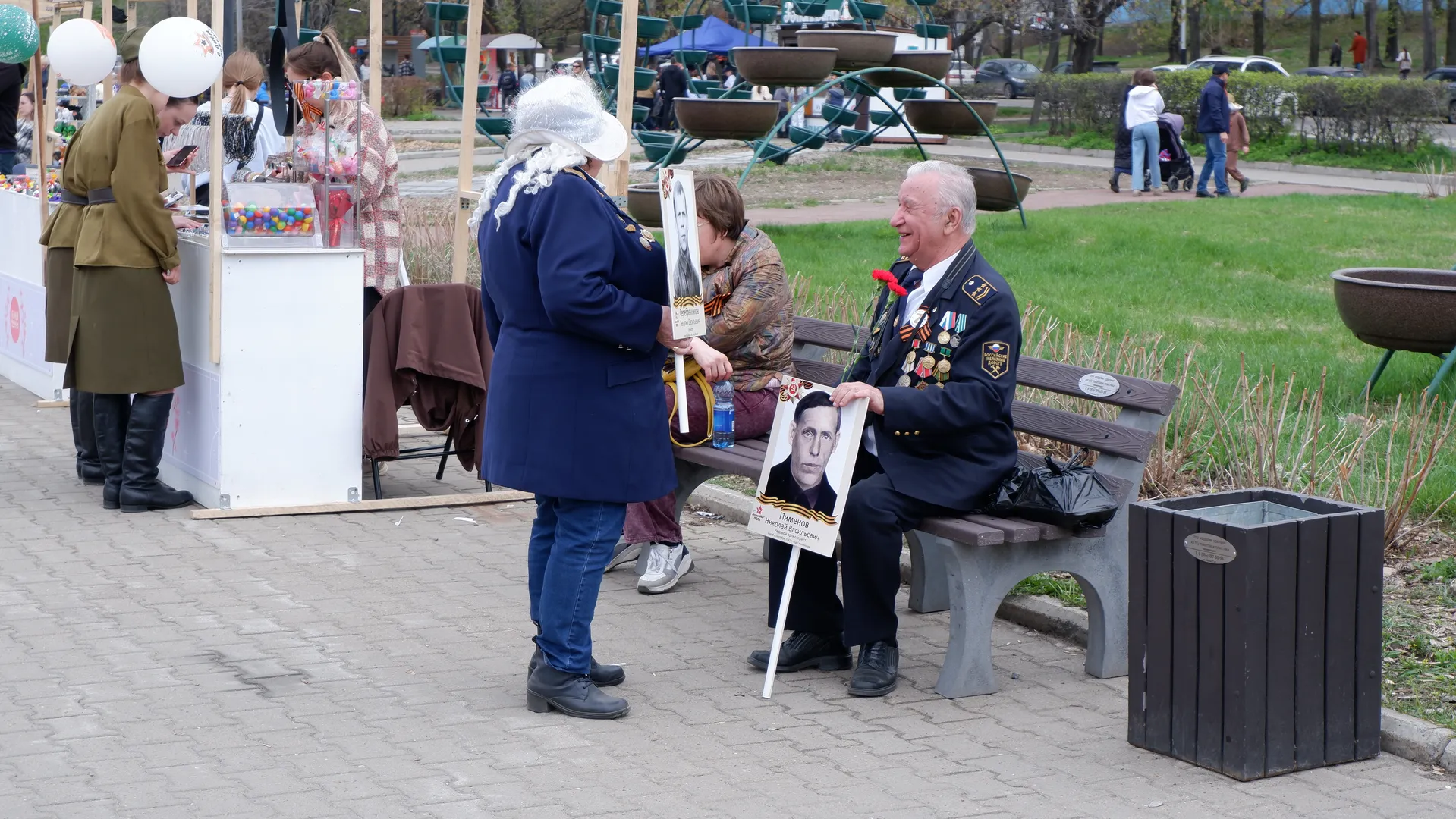 An elderly man in military uniform is sitting on a bench, with a woman in a blue coat standing next to him. There is a display board in front of the elderly man with photos and text. In the background, there are amusement rides and pedestrians.