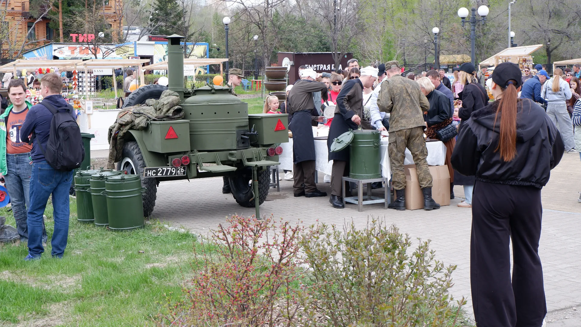 A green military vehicle is parked beside the crowd, covered with a blanket. People are gathered around tables and barrels, seemingly engaged in some activity. The background includes trees and buildings.