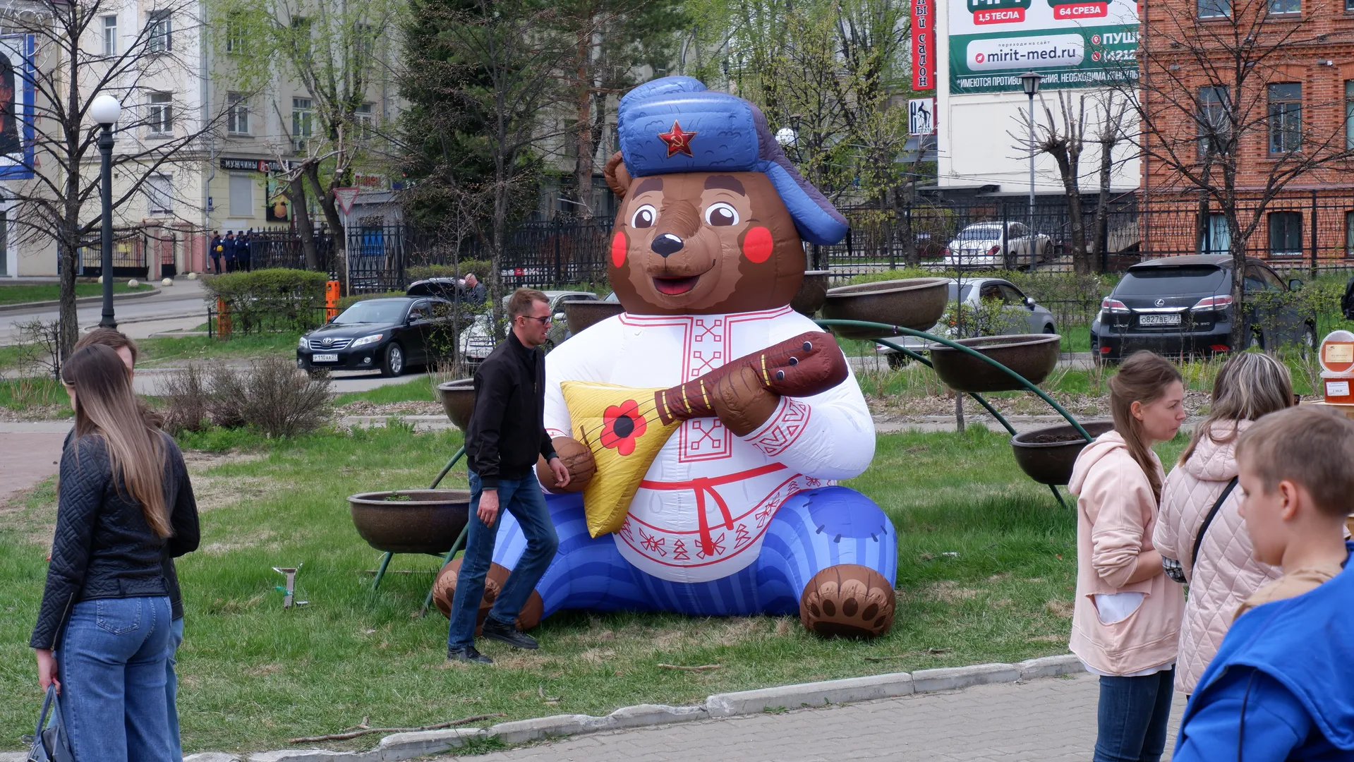 A giant inflatable bear sculpture is situated on the grass, wearing a blue hat with red stars and a white outfit. Several people are standing around the sculpture, observing. In the background, there are trees, cars, and buildings.