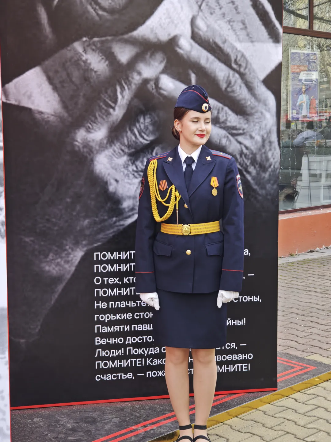 A woman in a dark blue uniform is standing in front of a poster, with yellow decorations and badges on the uniform. The poster shows an image of tightly clasped hands and accompanying Russian text. In the background, glass windows and an indoor scene can be seen.