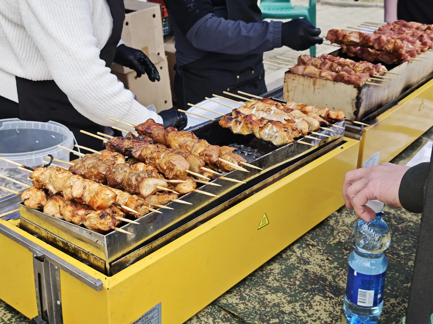 A yellow grill is holding several skewers of grilled meat, with the staff wearing black gloves to operate it. There is a bottle of blue-packaged water next to the grill. In the foreground, a hand is opening a bottle cap.