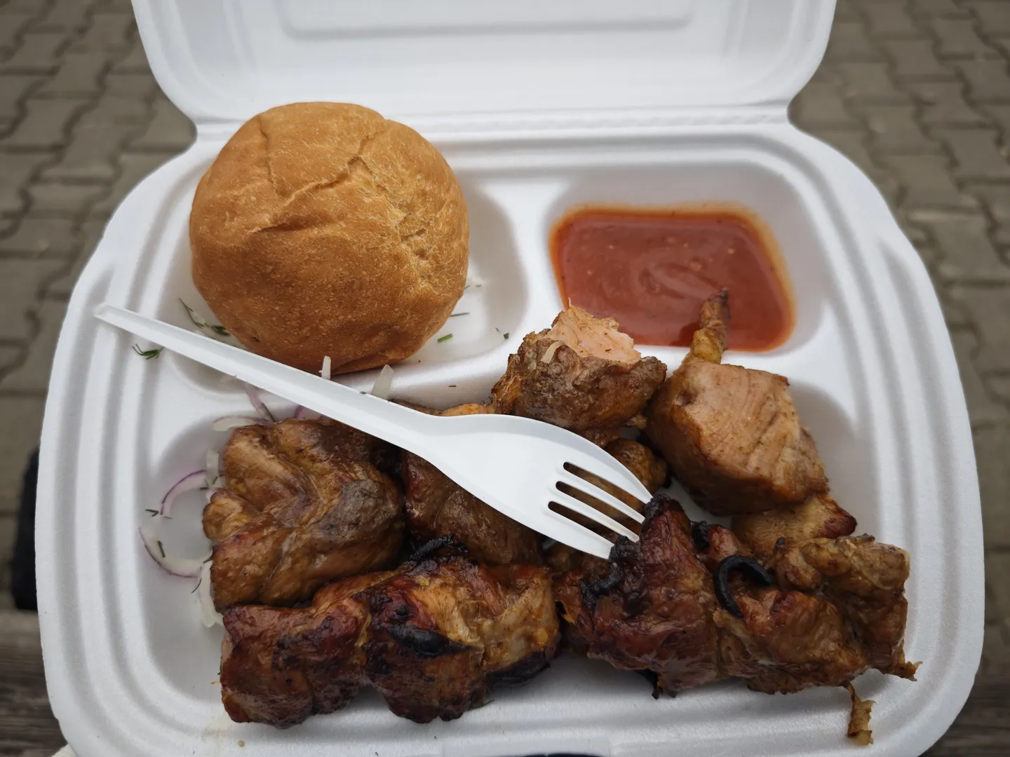A white foam food container holds grilled meat chunks, a small bread roll, and a portion of red sauce. A white plastic fork is placed on top of the container.