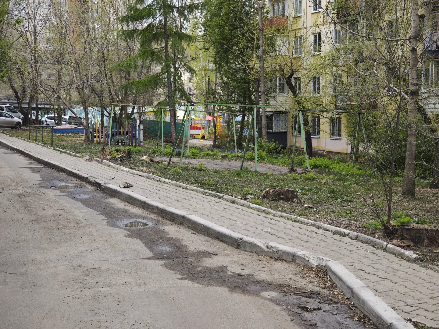 A brick-paved sidewalk is adjacent to a greenbelt, which has swings and other children’s playground equipment. In the background, parked cars and buildings can be seen. The sidewalk has traces of water.