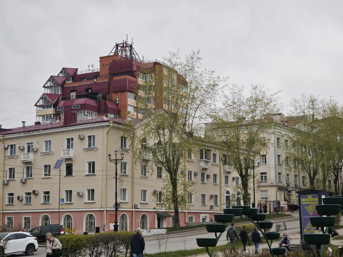 Multi-story buildings line both sides of the street, with a unified architectural style and beige exterior walls. Pedestrians are walking along the street, and trees are planted along the roadside. More buildings can be seen in the distance.