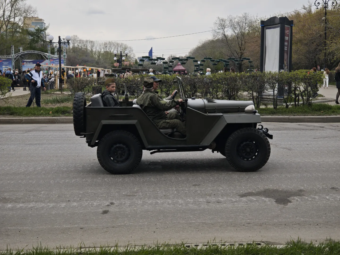 A military green jeep carries two individuals in military uniforms, driving on the road. In the background, there is a gathering of people, with surrounding greenery and streetlights. The sky is cloudy.
