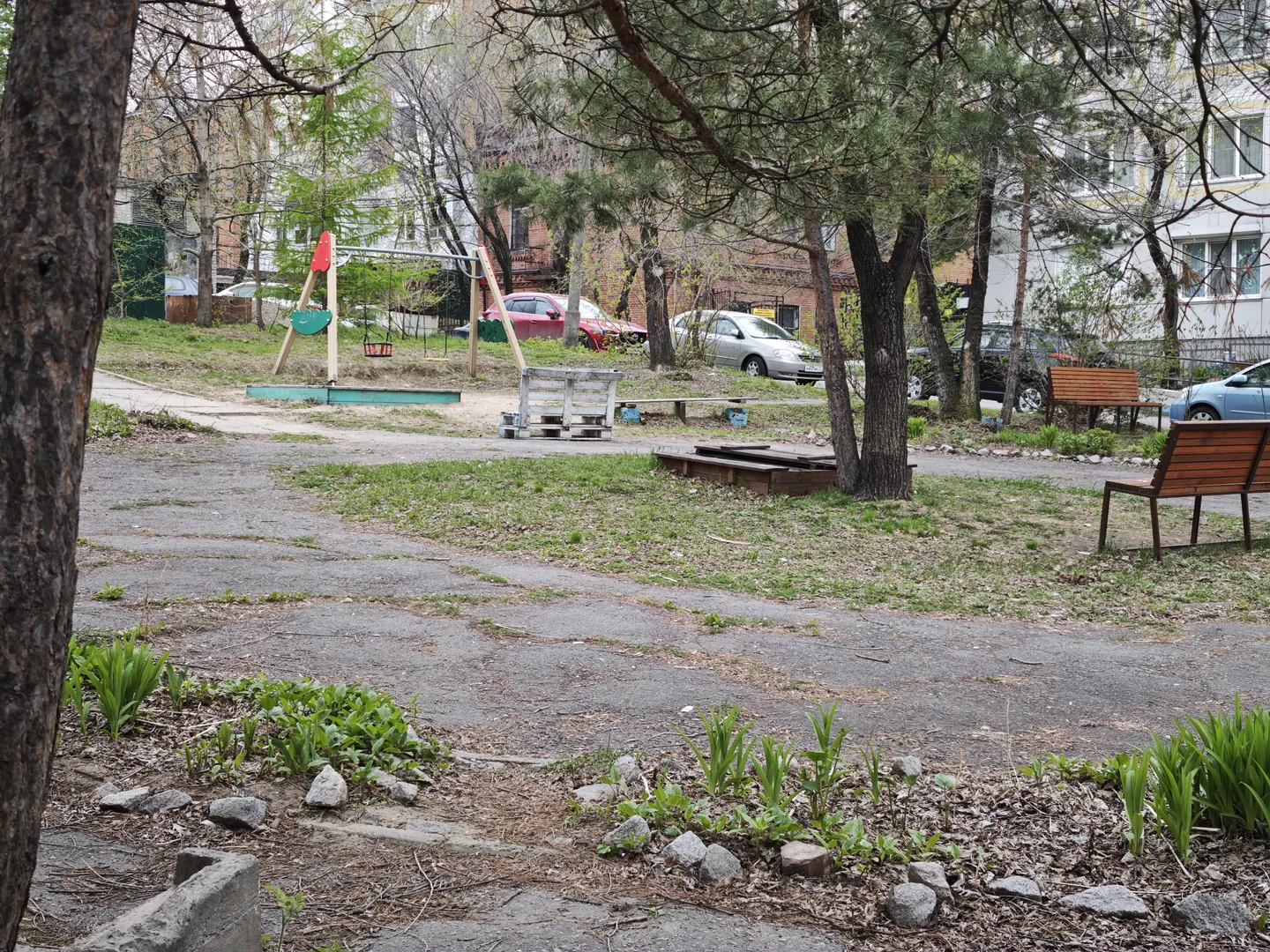 A wide open space, with the ground partially covered with grass and dirt, and part of it is paved with concrete. There are several wooden benches and children’s playground equipment. In the background, several cars are parked.