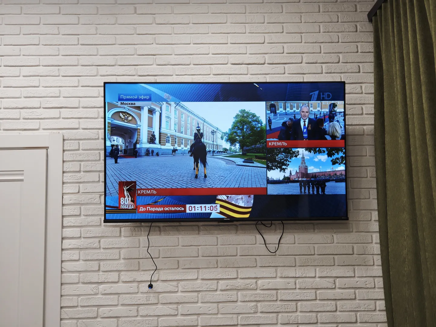 A TV is hanging on the wall, displaying street scenes and news reports from Moscow. The bottom left corner of the screen shows the word “КРЕМЛЬ,” and the top right corner shows the “1 HD” logo. There are wires connected below the TV.