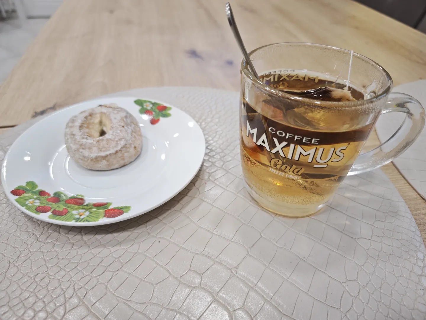 On a table, there is a transparent glass cup and a white plate. The glass cup contains tea, with the label “COFFEE MAXIMUS Gold FREEZE-DRIED” printed on it. On the plate, there is a donut dusted with powdered sugar. The surface of the table has an alligator skin texture.