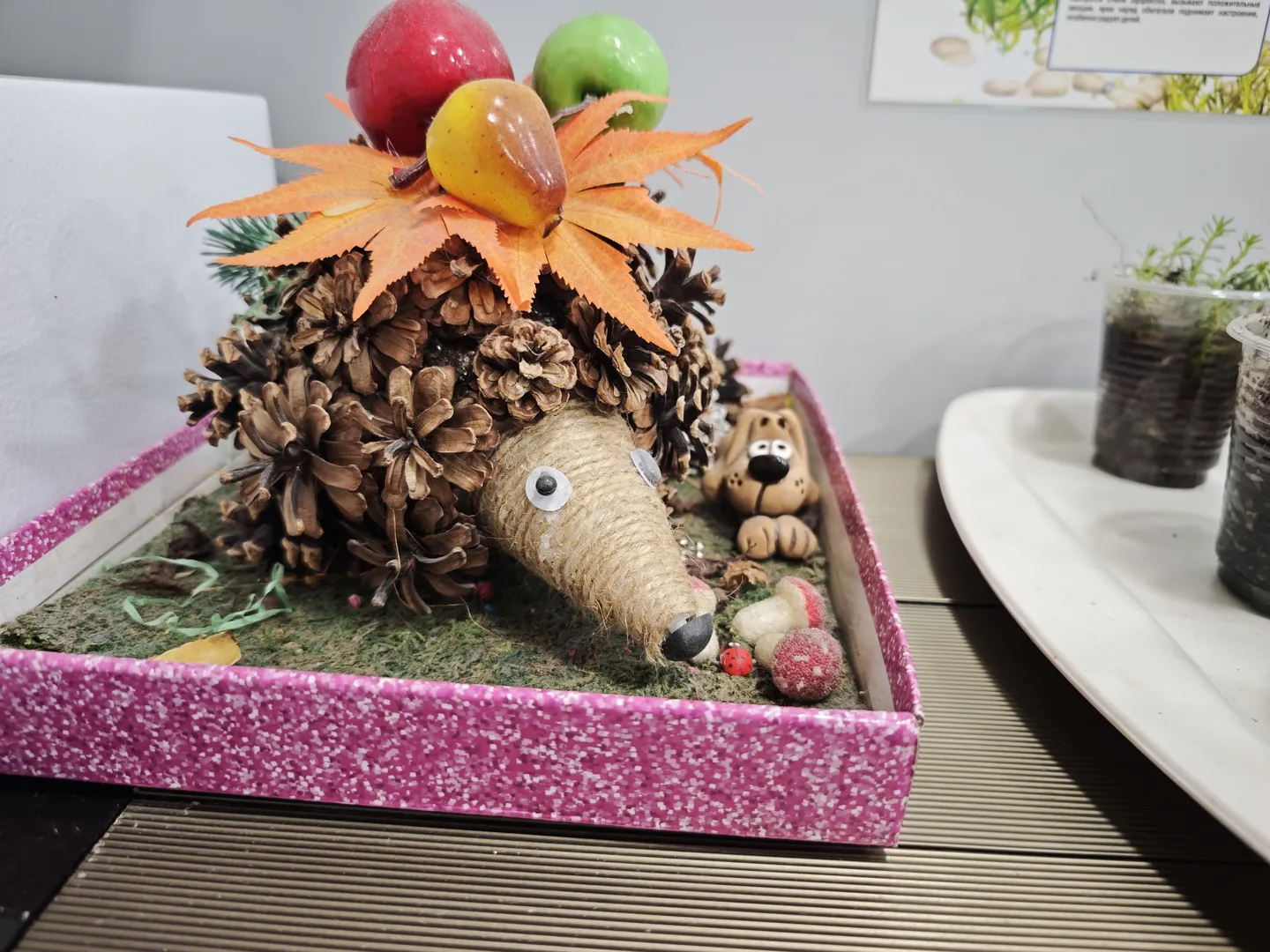 A handmade hedgehog model is placed inside a pink box. The hedgehog’s body is made of pinecones, its head is made of twine, and its eyes are small white balls. The top of the hedgehog has orange leaves and fruit decorations. There is also a small dog sculpture and a mushroom model next to it.