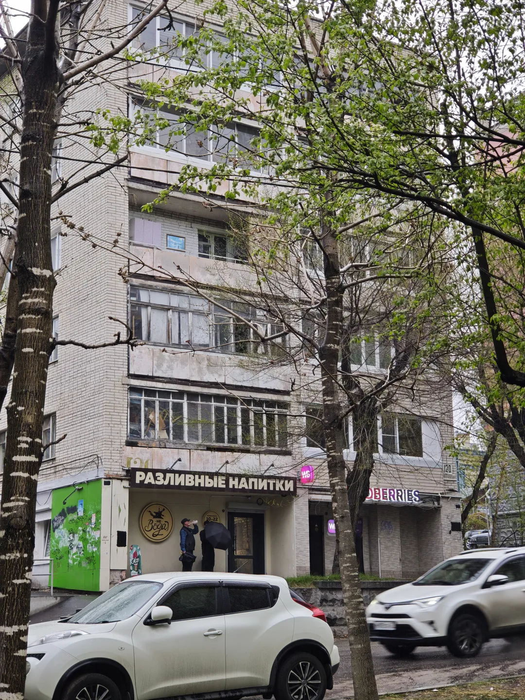 In front of a multi-story building, several cars are parked. On the ground floor of the building, there is a shop named “РАЗЛИВНЫЕ НАПИТКИ.” The tree branches partly obscure the building, and pedestrians and vehicles pass by on the street.
