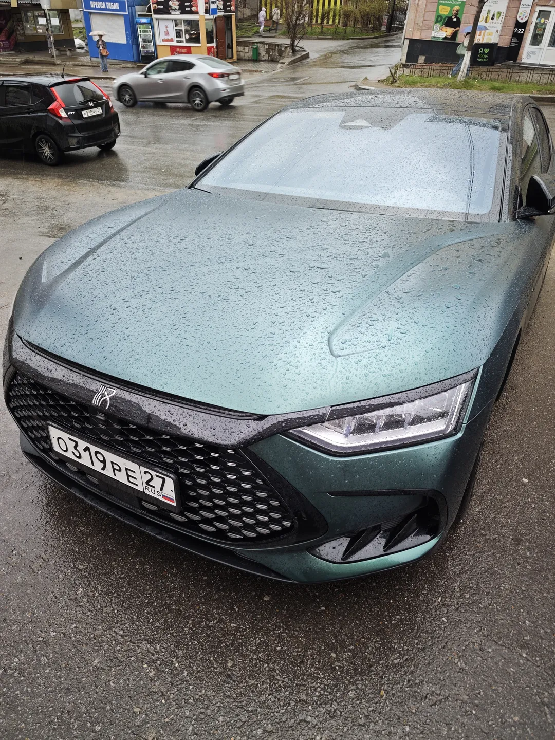 A dark green car is parked on a wet road surface, with raindrops on the car body. In the background, other vehicles and some buildings are visible.