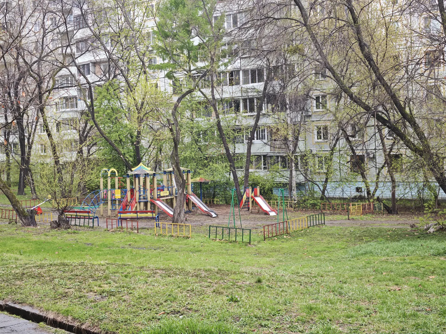A playground is located in front of multi-story residential buildings. The playground equipment includes a slide and climbing frame, surrounded by trees and green grass. The equipment is brightly colored, mainly red, yellow, and blue. The ground is covered with green grass, with some dirt and paths along the edges.