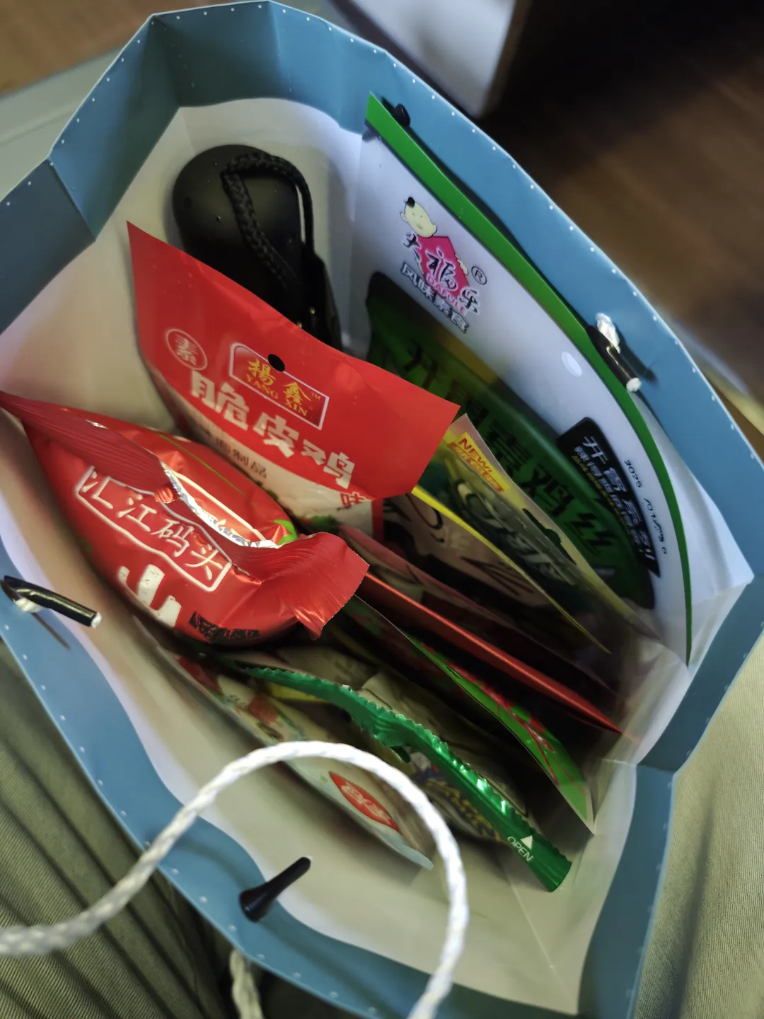 A blue paper bag contains various items, including food bags with red and green packaging and a small black object. The red packaging has the words “Yang Xin Crispy Chicken” printed on it, and the green packaging has the words “Appetizer Series.” There are also some other small packaged foods inside the bag.