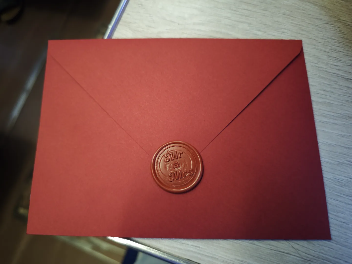 A red envelope is lying flat on a table, with a round wax seal in the center. The seal is engraved with the words “Mr & Mrs.” The envelope’s surface is smooth, and the background shows a light-colored wooden textured table. There are some blurry outlines of objects nearby.