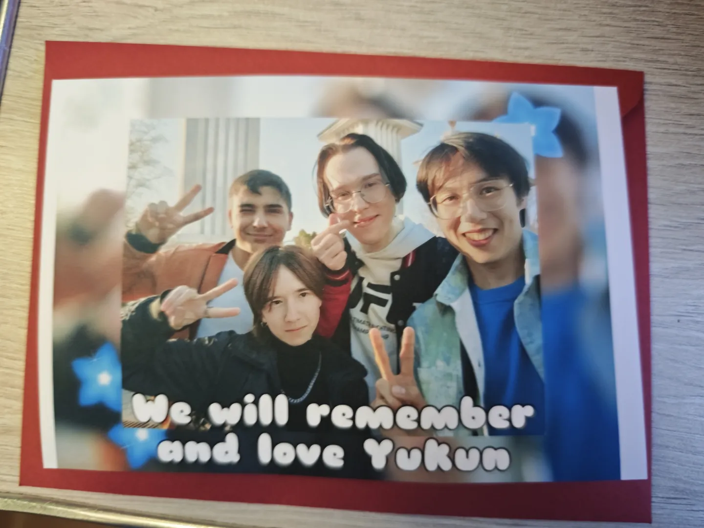 In a photo, four people are making victory gestures. The caption below the photo reads, “We will remember and love Yukun.” The edges of the photo are decorated with a red border and blue star patterns. In the background, parts of a building structure are visible.