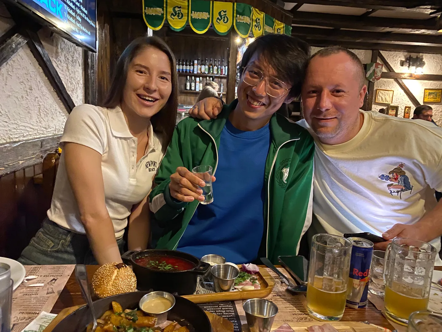 Three people are sitting at a dining table. The person in the center is wearing a green coat and a blue inner jacket, holding a glass of transparent drink. The person on the left is wearing a white short-sleeve shirt, and the person on the right is wearing a white T-shirt with a pattern. The table is set with food, drinks, and tableware, with a bottle of alcohol and decorative flags in the background.