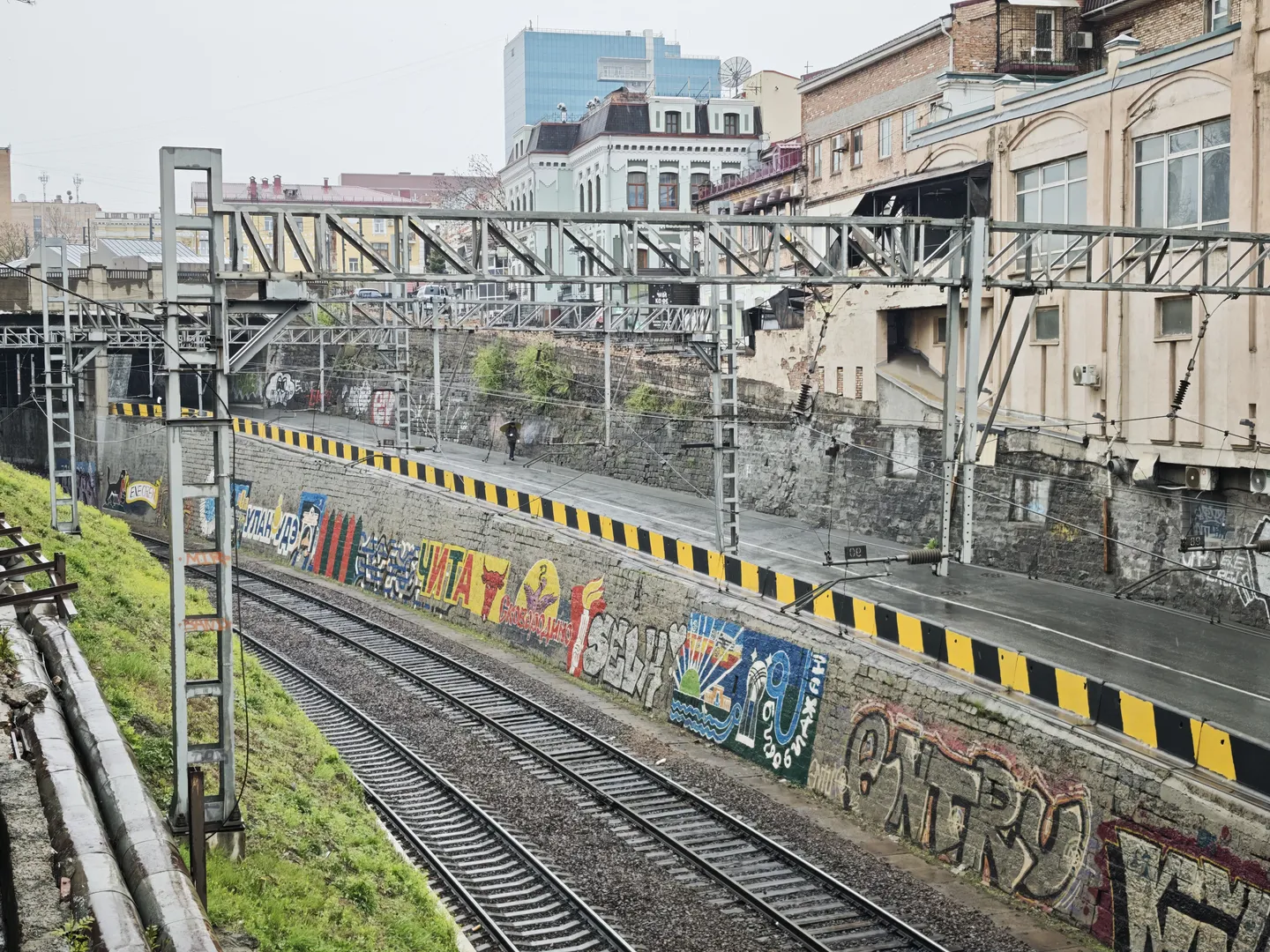 Next to the railroad tracks, there is a road with a metal support structure overhead. The wall beside the tracks is covered in colorful graffiti, including the word “ЧИТАТО.” In the distance, there are several high-rise buildings and low-rise residential blocks.