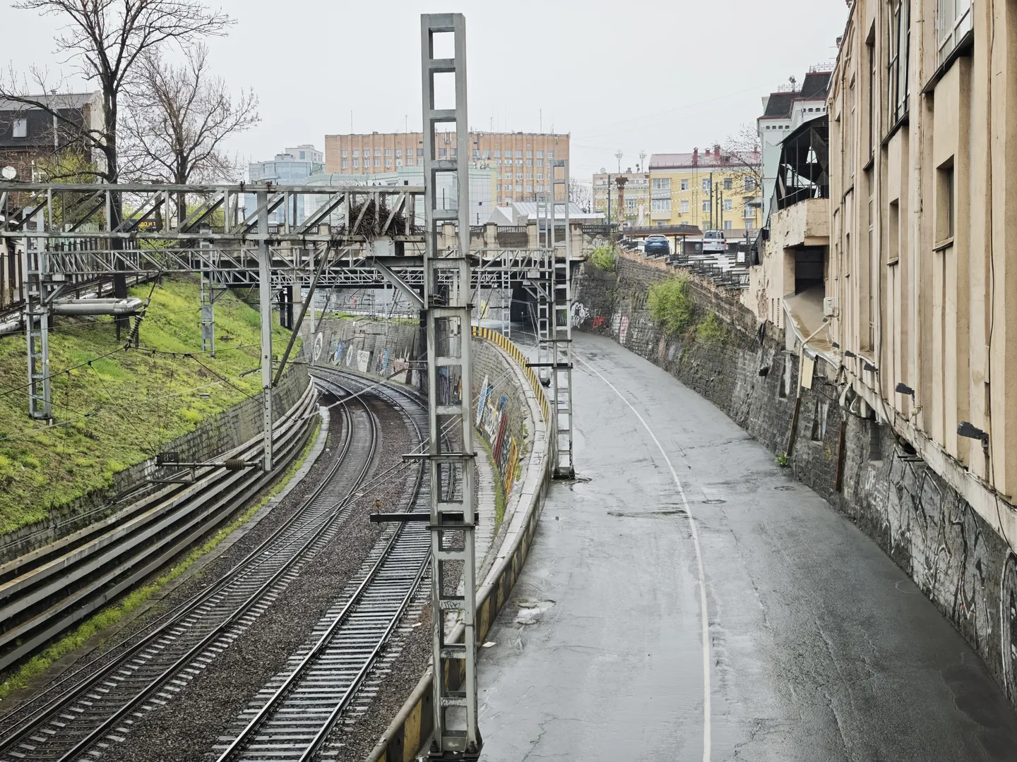 The railroad tracks stretch along a winding road, with a metal support structure above them. On one side of the tracks, there is green grass, while the other side features a wall with graffiti and buildings. Some high-rise buildings are visible in the background.