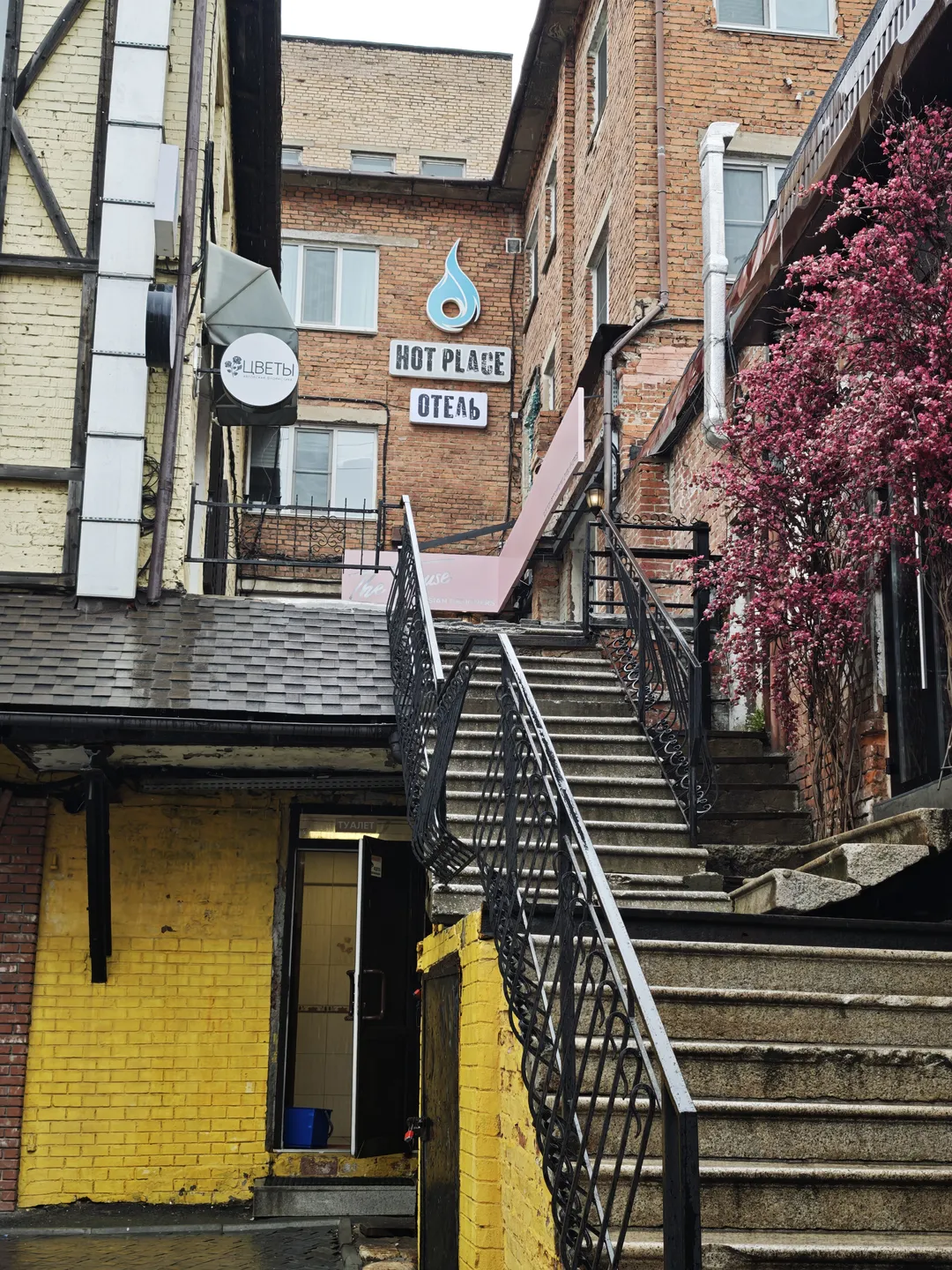 In front of a building with a yellow exterior, there is a staircase with black metal railings. The building displays a sign reading “HOT PLACE ОТЕЛЬ,” and next to it is a tree with pink flowers. The staircase leads to an entrance on the second floor.