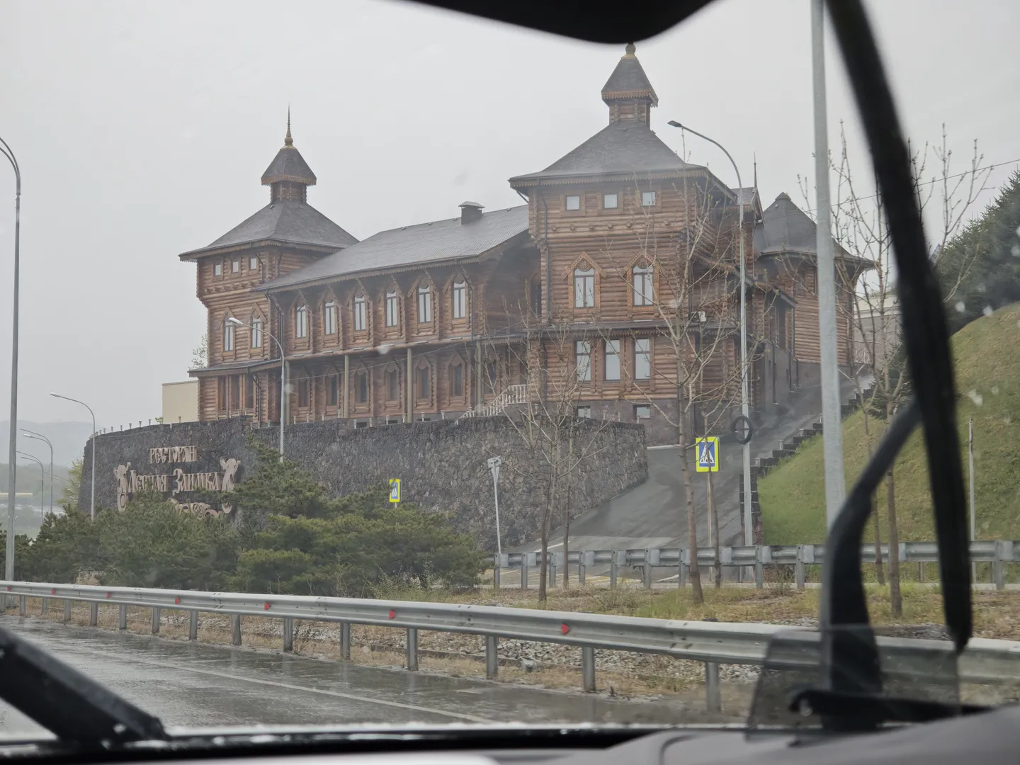 From inside a vehicle, we see a large wooden building with multiple windows and a pointed tower. In front of the building, there is a railing and several trees. The sky looks overcast, and part of the car’s windshield wiper is obscuring the view.