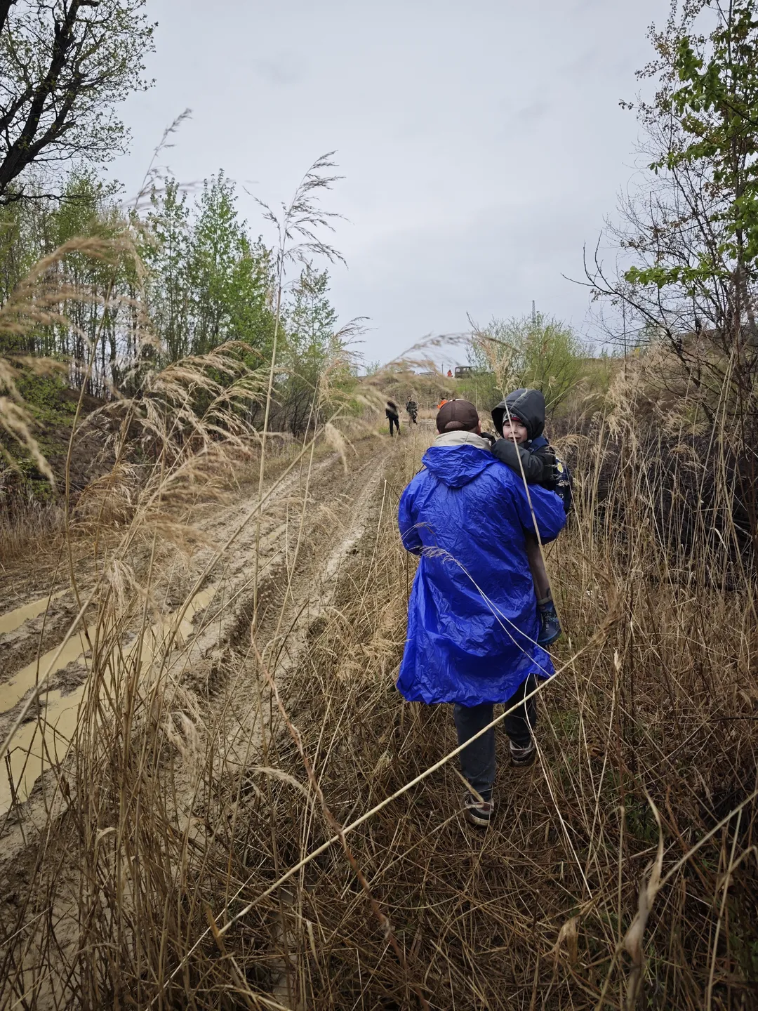Two people in blue raincoats walk across a grassy field surrounded by tall hay. In the distance, several people are also moving around the field, with trees and a vehicle in the background. The sky is overcast.