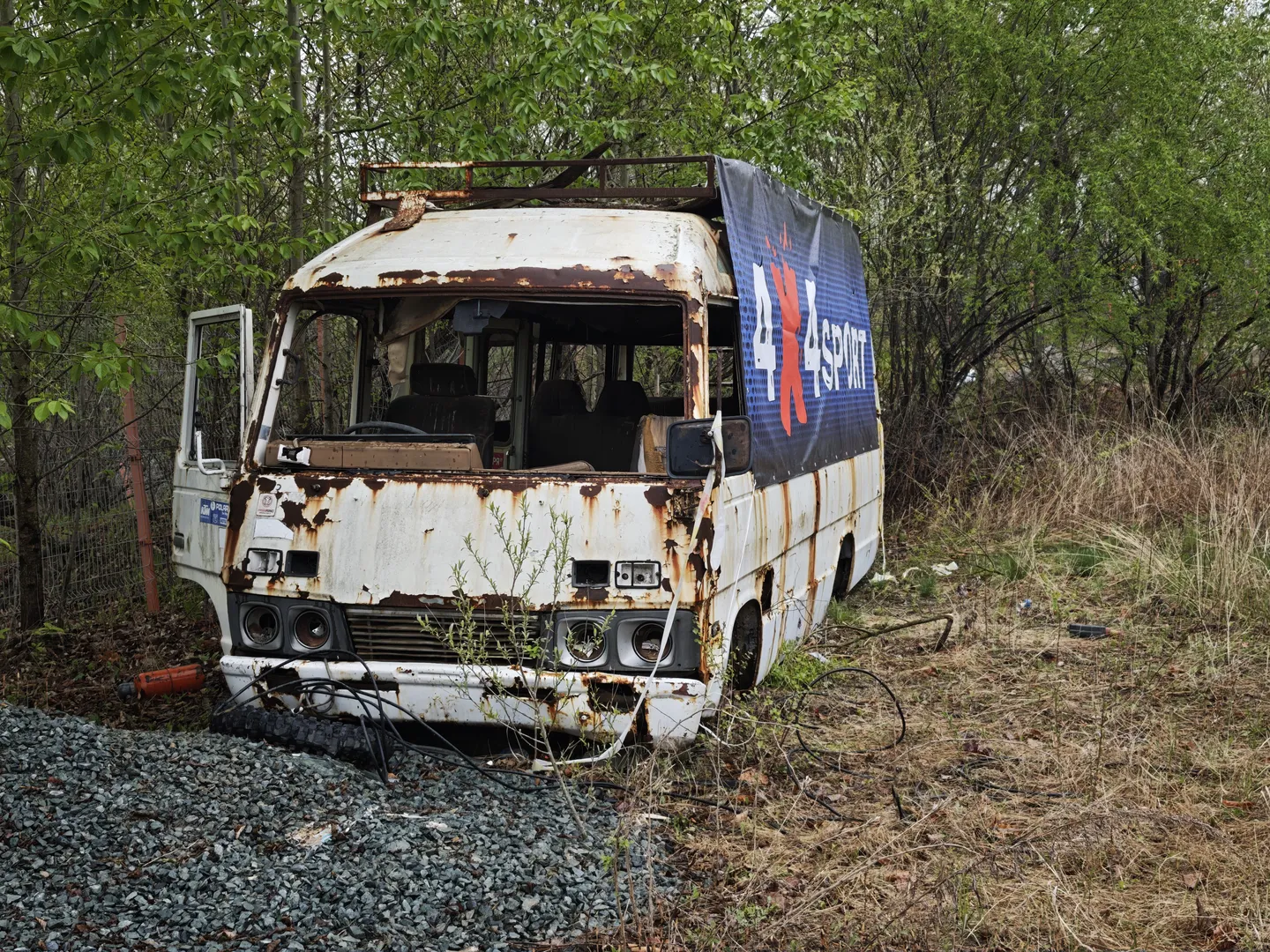 An old white bus is parked in a wooded area, with heavy rust on the body. The side of the bus displays an advertisement for “4x4sport,” and some windows are broken. The ground is covered with dry grass and gravel.