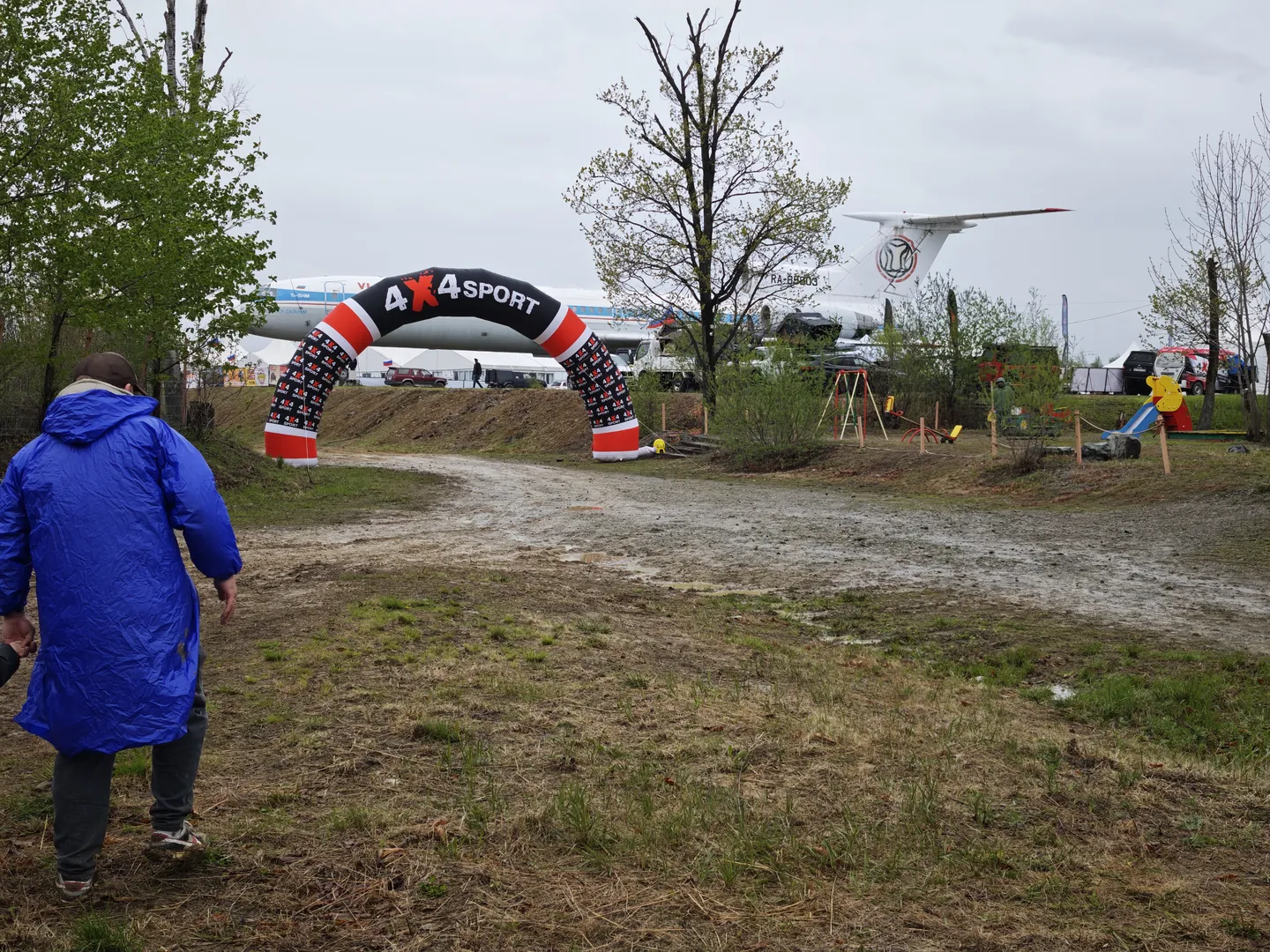 A person in a blue raincoat stands on muddy ground in front of an arch labeled “4x4SPORT.” In the background, a large airplane is parked, with tents and amusement facilities nearby. The ground is slippery with visible tire tracks.
