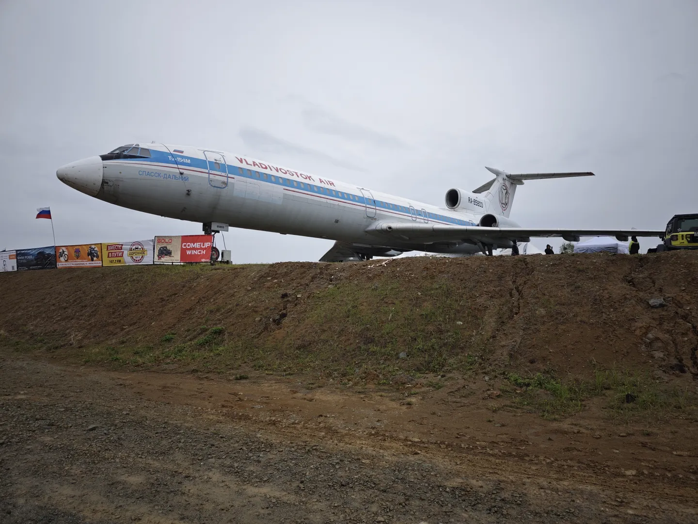 A large airplane is parked on a dirt slope, with the words “VLADIVOSTOK AIR” on its fuselage. In front of the airplane, there are flags and an advertisement, with trees visible in the background. The ground is flat and covered with dirt.