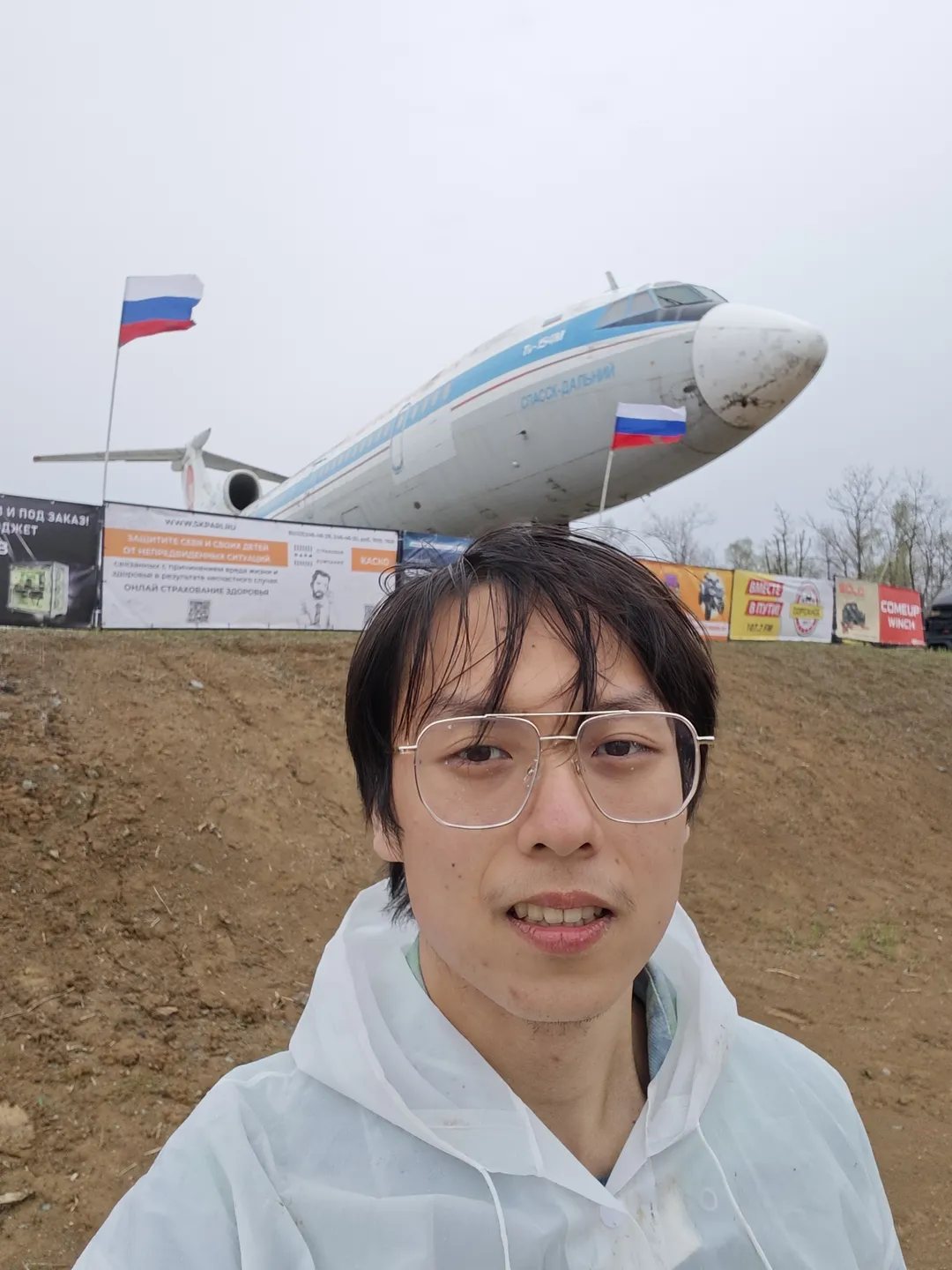 A person in a white coat stands outdoors, with a large airplane in the background displaying the words “VLADIVOSTOK AIR” on its fuselage. Flags and advertisements are in front of the airplane, with an overcast sky. The ground is flat and covered with dirt.
