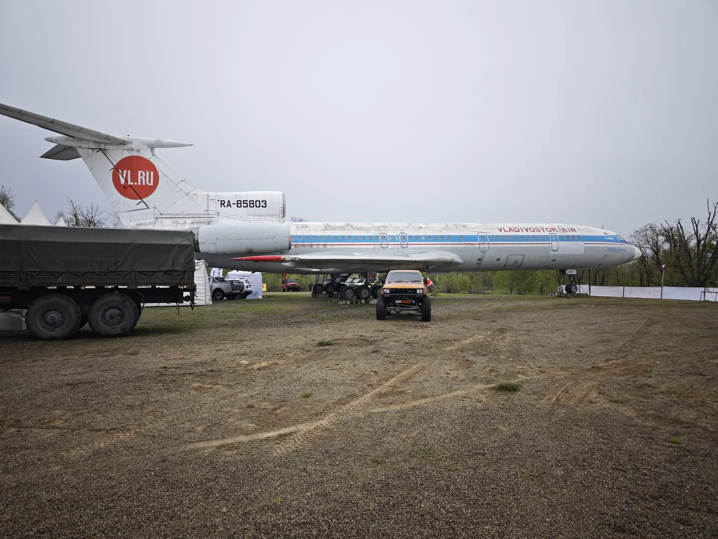 A large airplane is parked on an open area, with the tail marked “VL.RU.” In front of the airplane, a jeep and a truck are parked, with trees and tents in the background. The ground is flat and covered with dirt.
