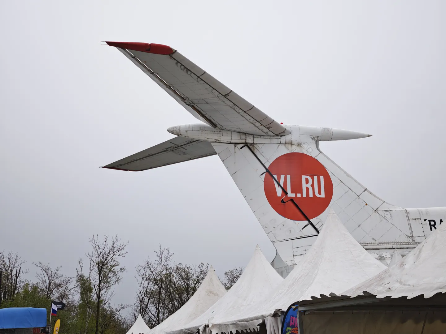 The tail of the airplane features a red circular logo with the text “VL.RU.” The tail and fuselage of the airplane are visible, with a gray sky in the background. In the foreground, a white tent top can be seen.