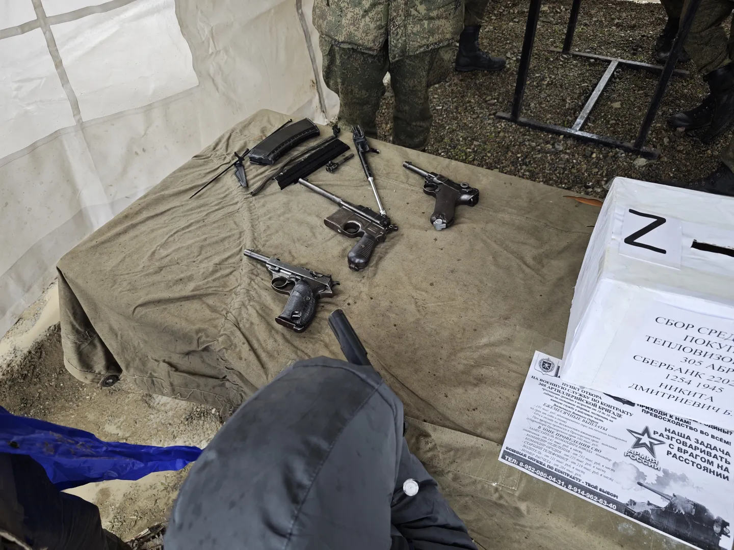 On a table, several handguns and accessories are laid out, with a person in camouflage standing nearby. The table is covered with light-colored fabric, and a piece of paper with text and patterns is on the right side. The background is an indoor setting.