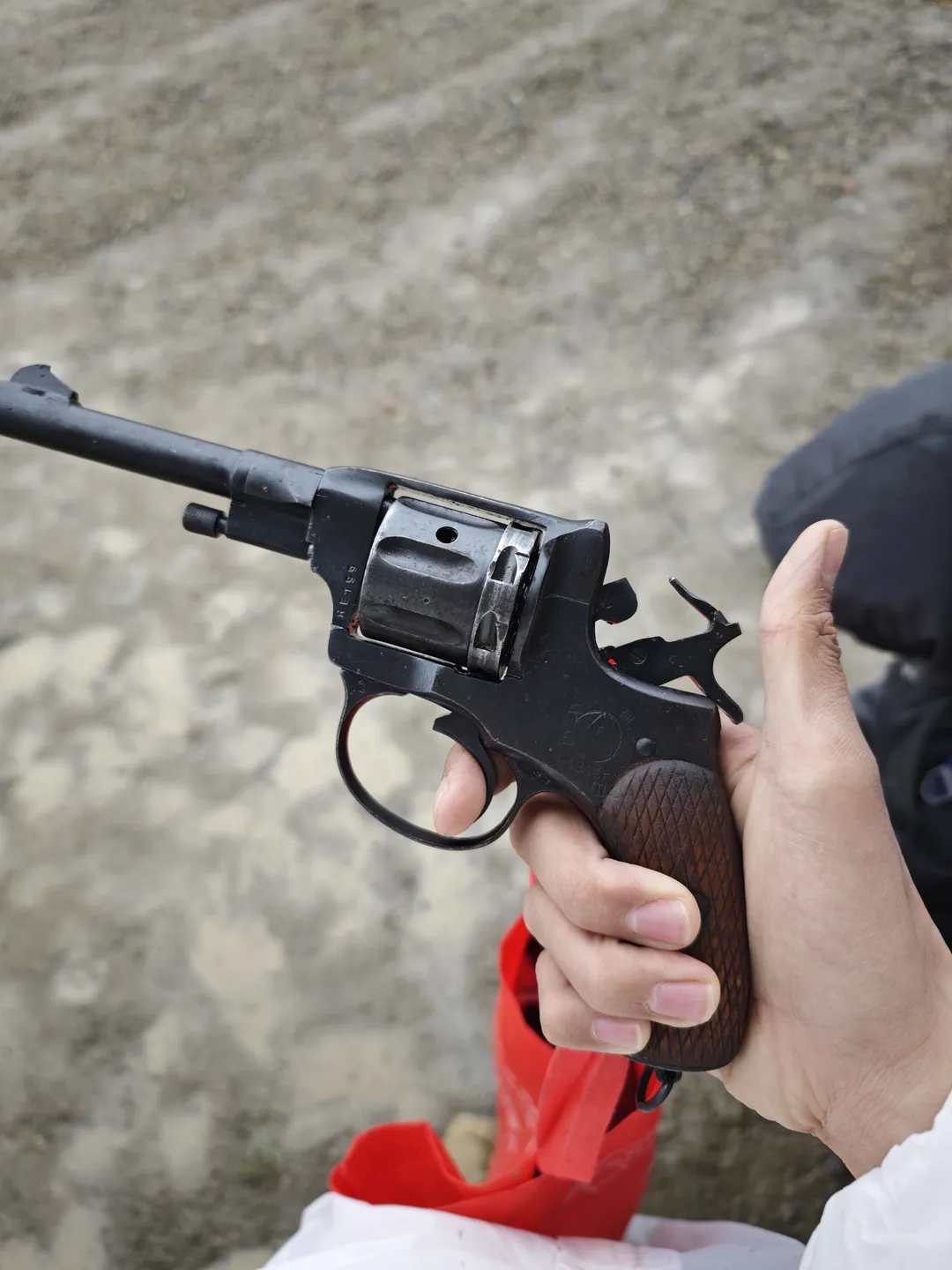 A hand grips a black handgun, with a wooden-textured grip. The background is a gray floor, and a person in dark clothing stands beside. The details of the handgun are clearly visible.
