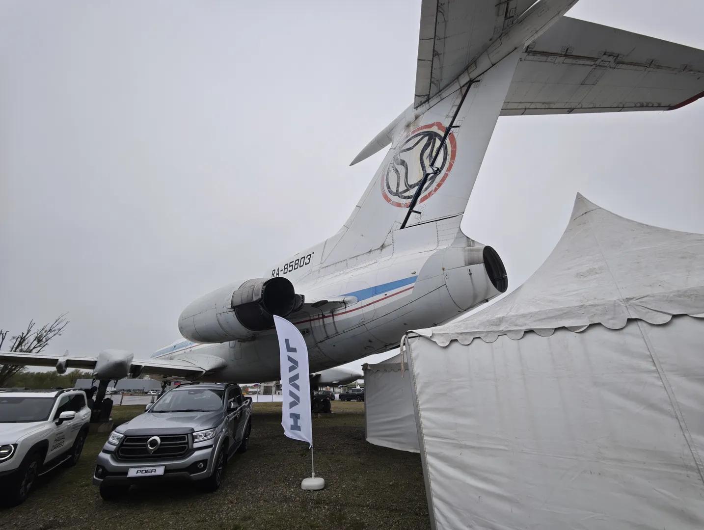 A large airplane is parked next to a white tent, with the number “RA-85803” on its fuselage. The tail of the airplane features a unique logo, with overcast skies in the background. Several cars are parked in the foreground.