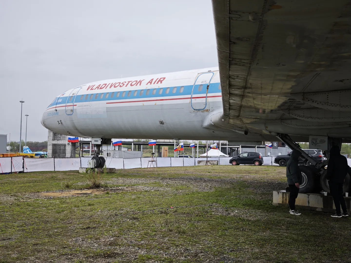 A large airplane, marked “VLADIVOSTOK AIR,” is parked on a grassy field. The airplane’s fuselage is white with blue and red stripes. Several people are standing near the airplane, with fences and flags visible in the background.