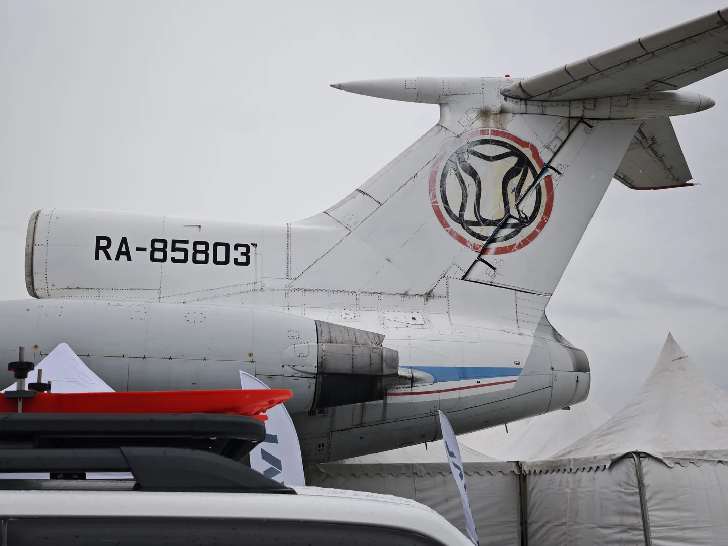 The airplane’s tail shows the number “RA-85803,” and its tail wing features a unique logo. The airplane is parked next to a white tent, with overcast skies in the background. The airplane’s engine and tail wing details are clear.