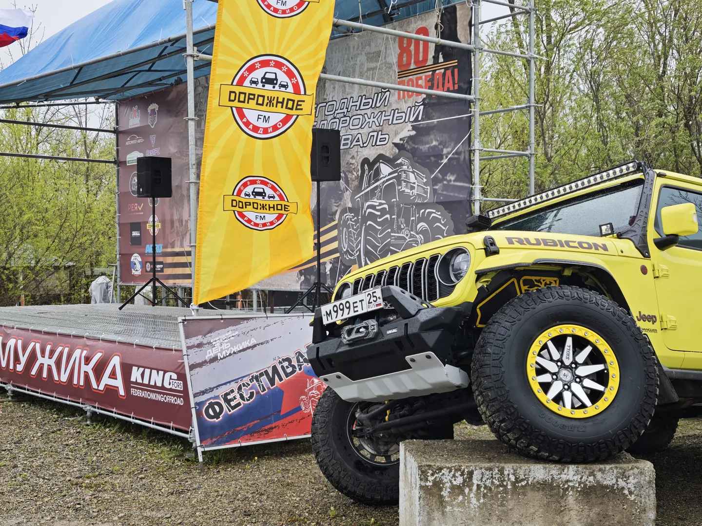 A yellow Jeep Rubicon is parked in front of a stage, with large wheels and a robust body. Several banners and slogans are displayed on the stage, with trees in the background. One of the banners reads “ДОРОЖНОЕ FM.”