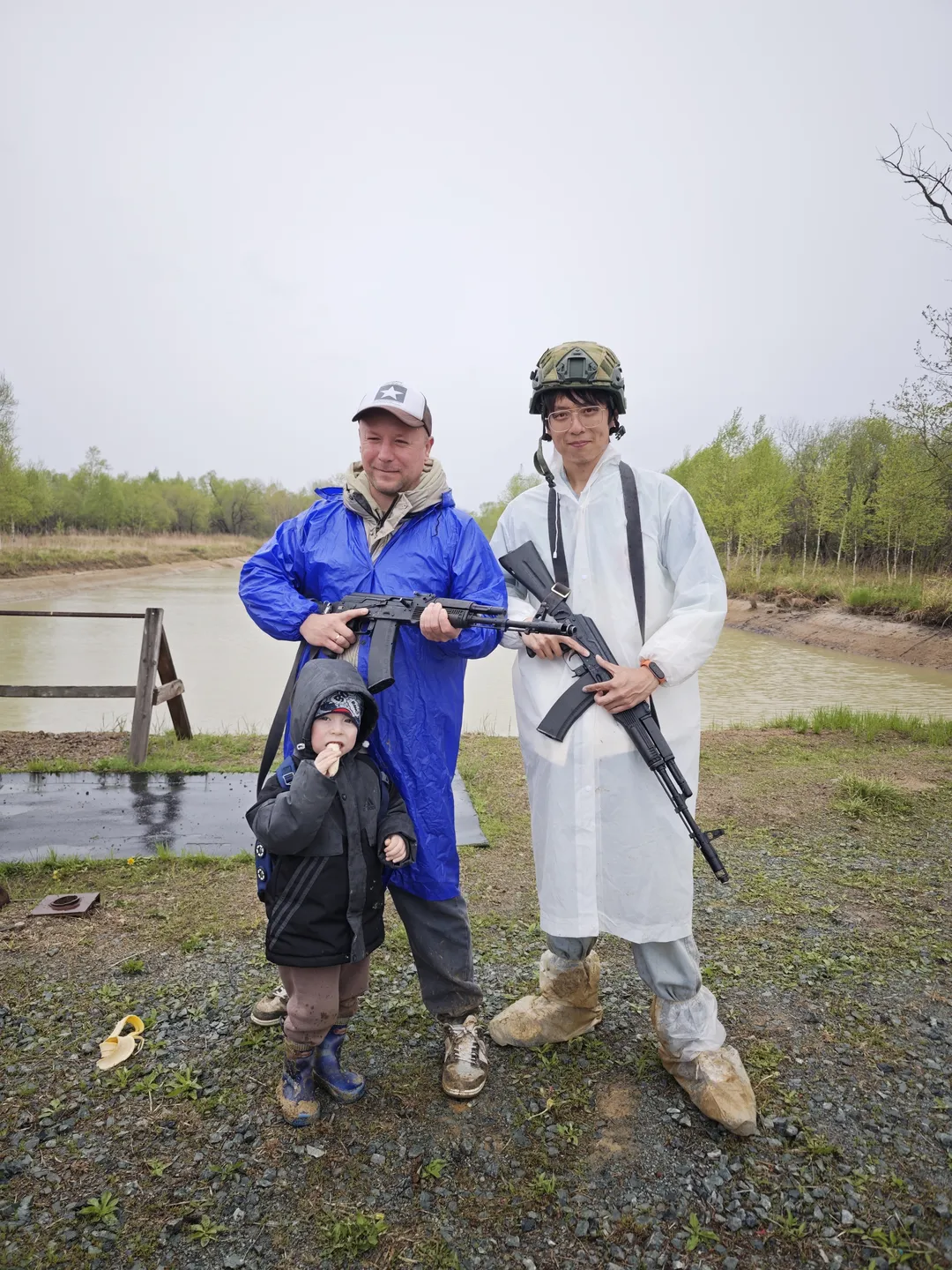 Two people and a child are standing together, each holding a gun. The person on the left is wearing a blue jacket, while the person on the right is wearing a white jacket and a helmet. The background features a river and trees.