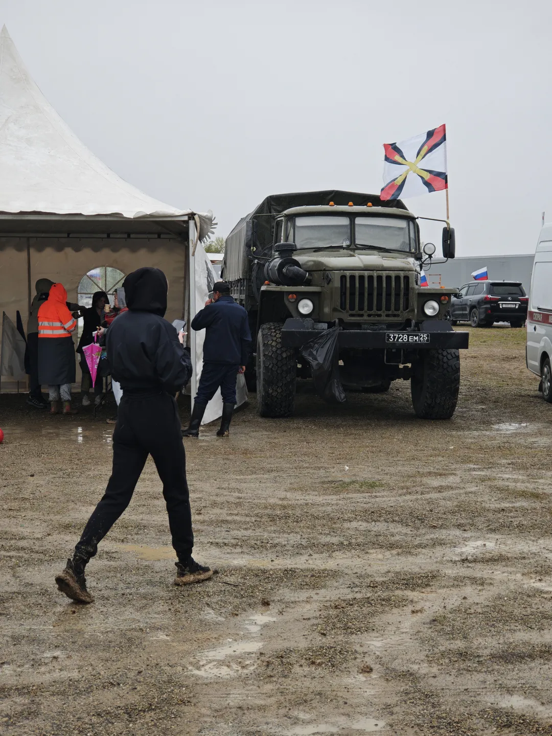 A military truck is parked next to a white tent, with a flag attached to its roof. Several people are moving between the truck and the tent, with the ground covered in dirt. Other vehicles and buildings can be seen in the background.