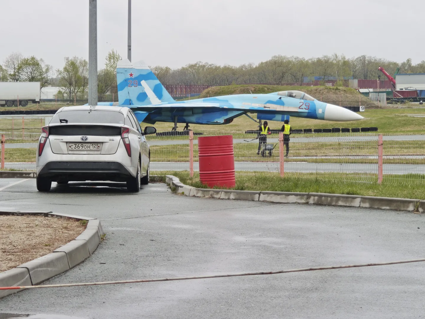 A white car is parked on the roadside, with a blue camouflage fighter jet in front. The jet is numbered “29,” with a grassy field and trees in the background. Two workers are seen working next to the jet.