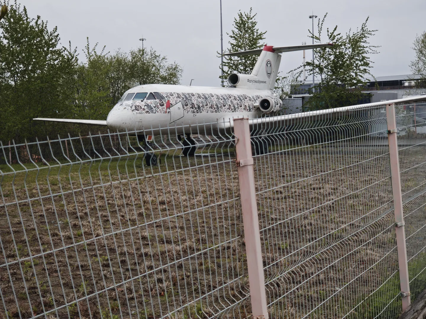 An airplane is parked on a grassy field, with patterns and the text “#допускасборщиков” on its fuselage. There are trees and a metal fence around the airplane. Some buildings are visible in the background.