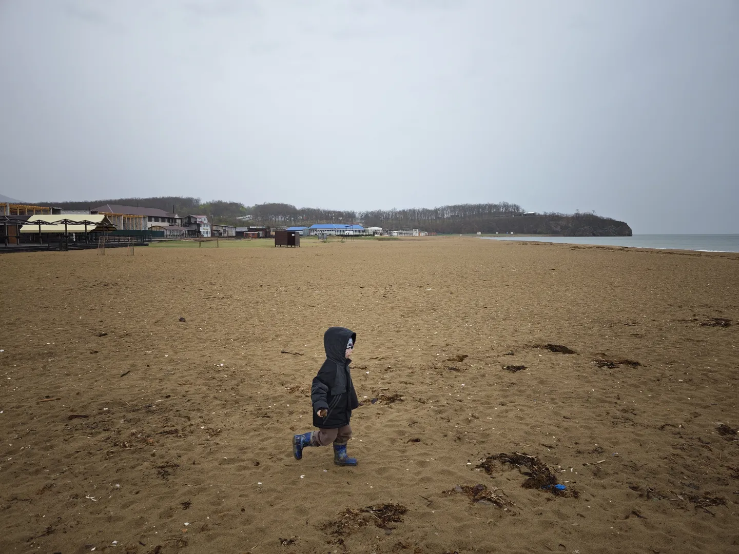 A child in a black coat and blue boots is running on a beach. The beach is vast, with buildings and trees in the distance. The sky is overcast.