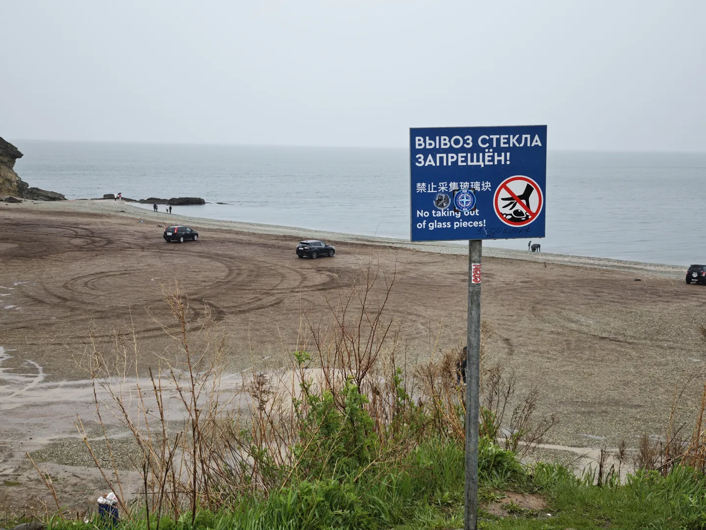 A sign stands on the beach, reading “ВЫВОЗ СТЕКЛА ЗАПРЕЩЁН!” along with “禁止采集玻璃块” and “No taking out of glass pieces!” Several vehicles are parked on the beach, with the ocean in the distance.