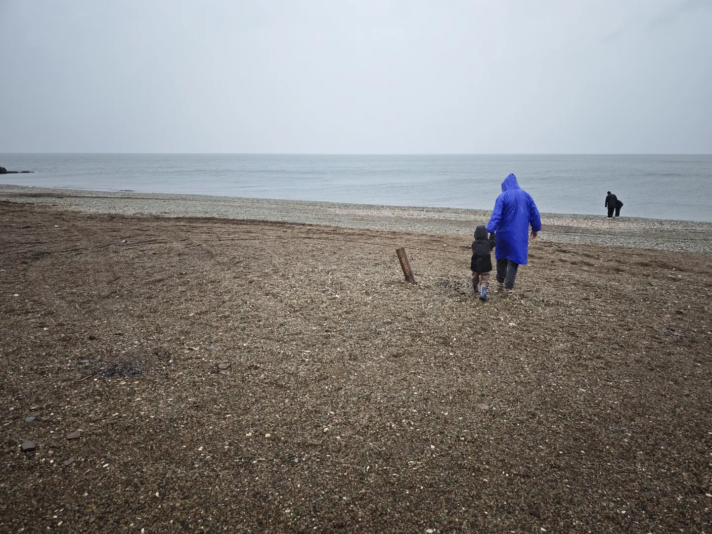Two people are walking on the beach, one wearing a blue raincoat and the other in dark clothing. The beach is covered with small pebbles, and the calm sea is visible in the distance. Another person is standing farther away.