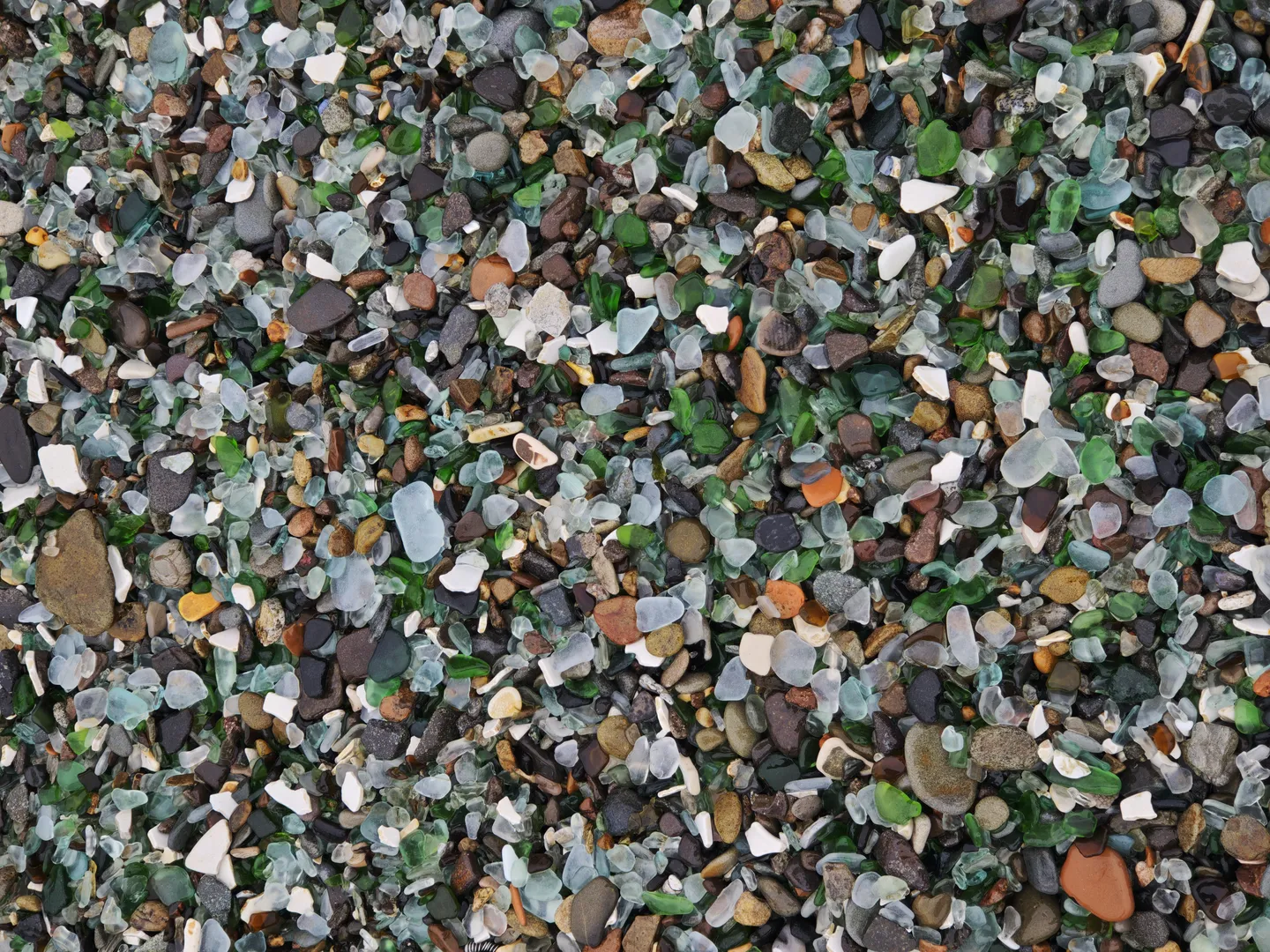 The beach is covered with various colored glass fragments and pebbles, including green, blue, white, and brown. The glass fragments are irregular in shape and vary in size, creating a colorful appearance.