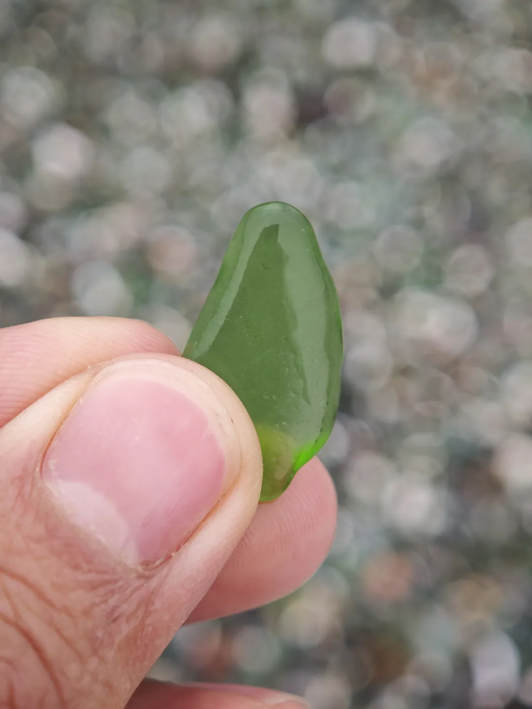 A hand holds a green glass fragment, with a blurred background. The glass is oval-shaped and smooth to the touch, with fingers gripping the edges of the fragment.