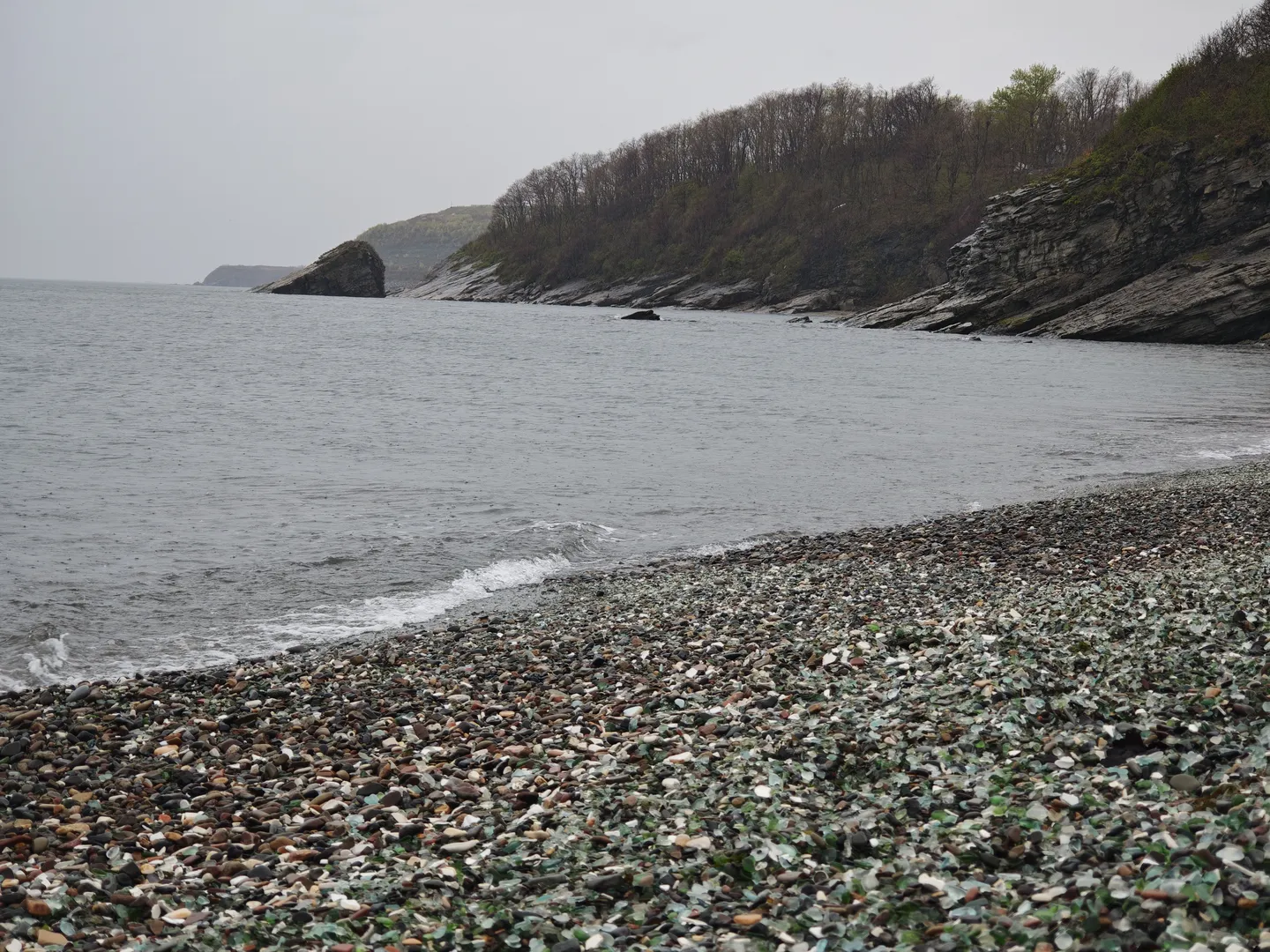 A coastline with a beach covered in glass fragments and pebbles. The waves of the sea are gently lapping against the shore, forming white foam. In the distance, there are hills covered with vegetation.